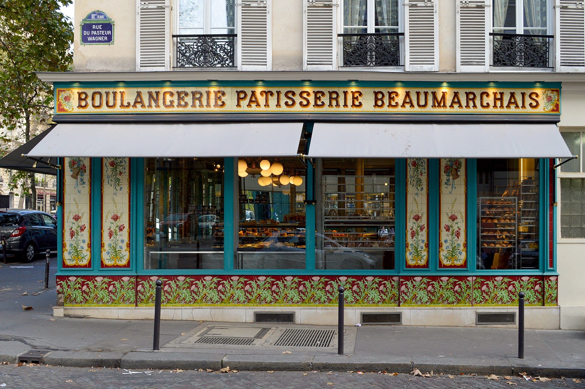 Tiled shopfront of a Paris boulangerie-patisserie with floral panels, a striped awning and trays of bread in the window