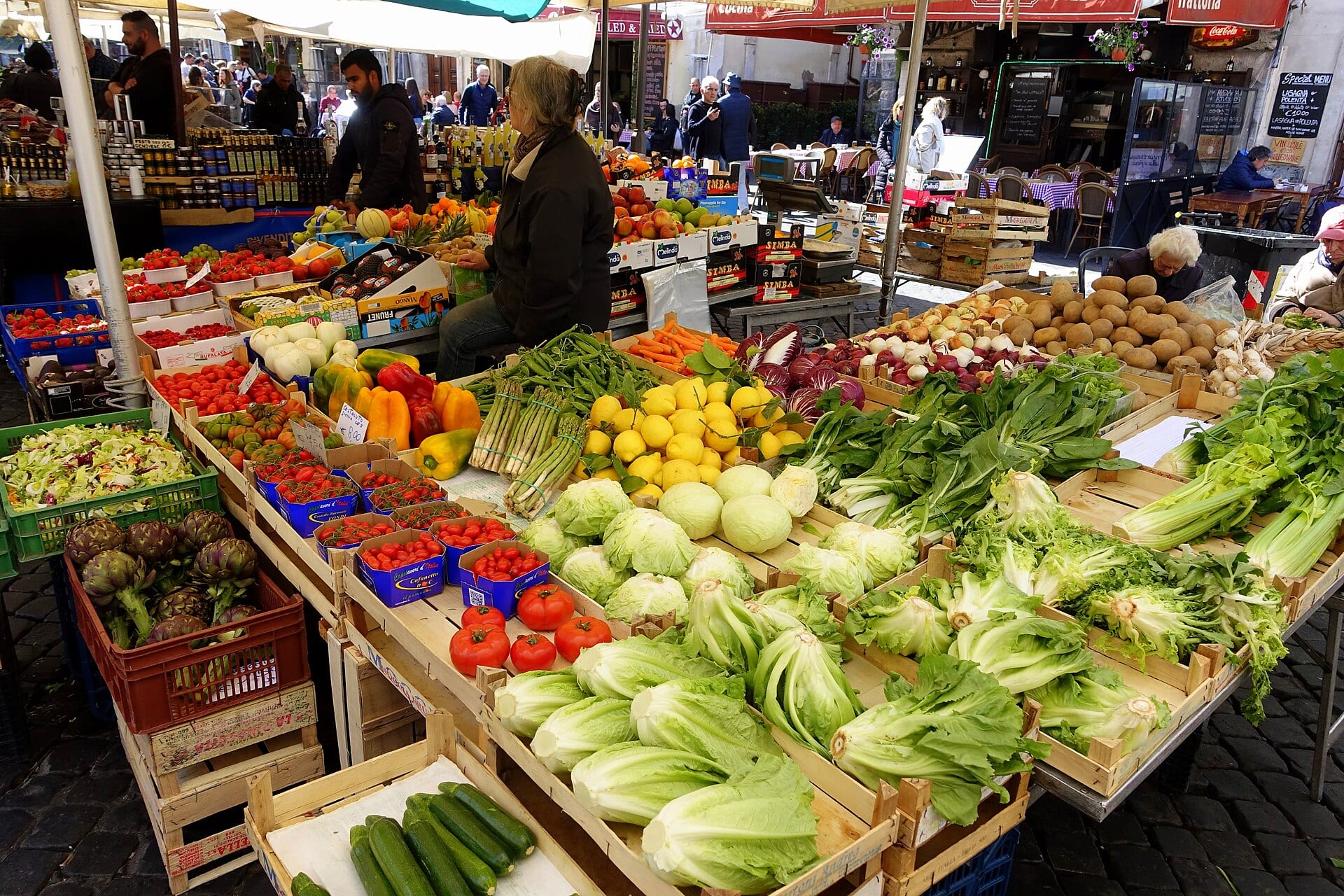A produce stall at a Rome open-air market with artichokes, tomatoes, lemons, asparagus and chicory in wooden crates