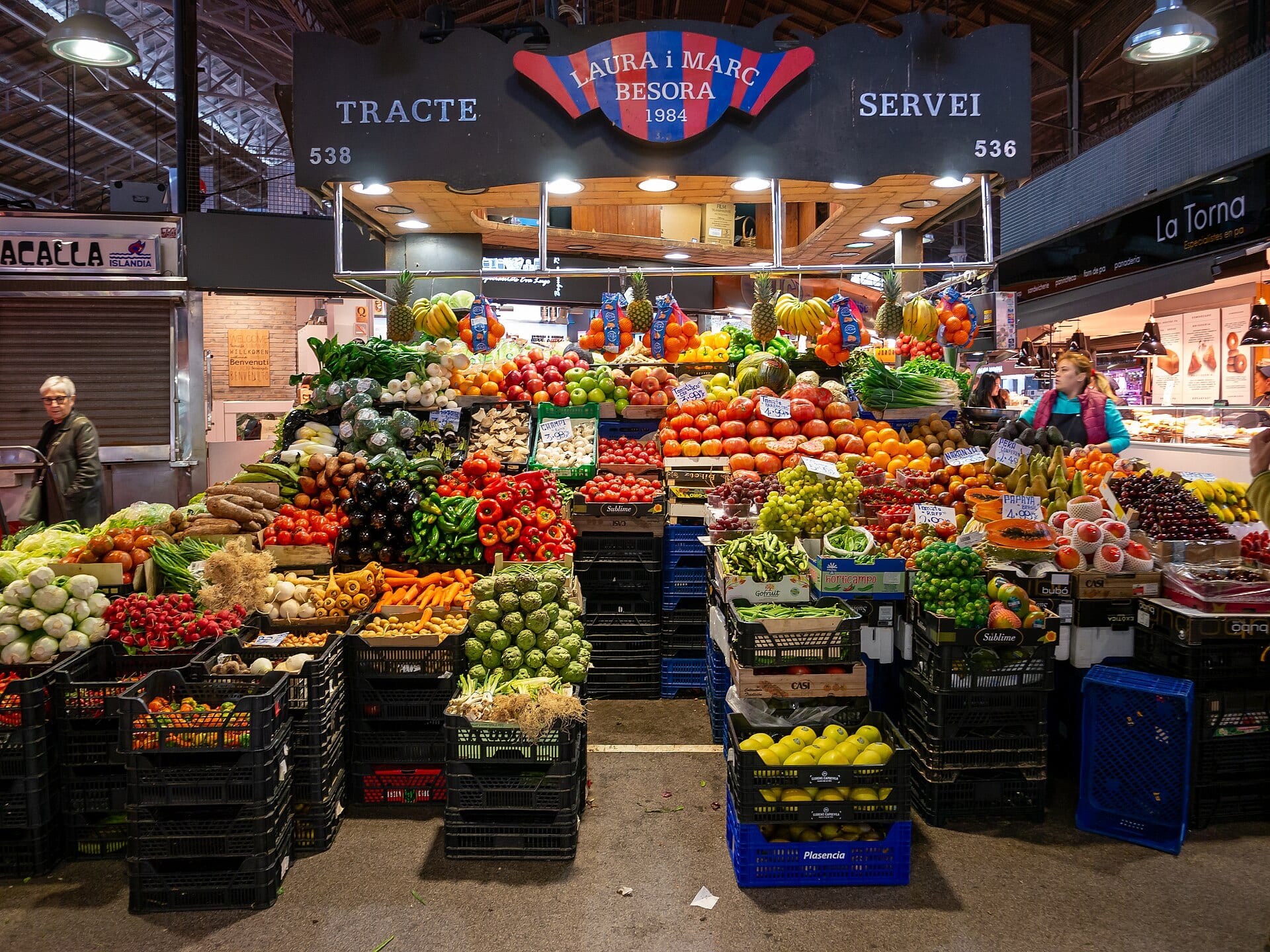 A heaped fruit and vegetable stall inside Barcelona's La Boqueria market under a hand-lettered sign